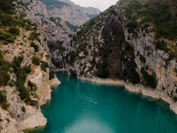 A serene river flows between rugged cliffs covered with lush green vegetation. The water is a vivid turquoise, creating a striking contrast with the earthy tones of the surrounding rocks. In the middle of the river, a lone kayak or small boat is visible, adding a sense of scale to the landscape. The scene is framed by distant mountains, under an overcast sky, enhancing the natural beauty of the setting.