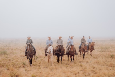 A group of six people wearing cowboy hats and casual attire are riding horses across an open, expansive field of dry grass. The sky appears overcast with a layer of fog creating a soft, muted backdrop.