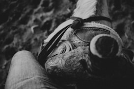 A close-up black and white image capturing a detailed view of a leather horse saddle with intricate patterns. A part of a rider's leg in denim is visible, suggesting they are seated on the saddle. The texture of the horse's mane is slightly visible towards the top.