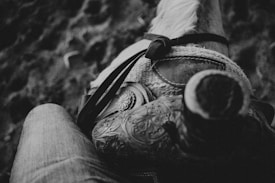 A close-up black and white image capturing a detailed view of a leather horse saddle with intricate patterns. A part of a rider's leg in denim is visible, suggesting they are seated on the saddle. The texture of the horse's mane is slightly visible towards the top.