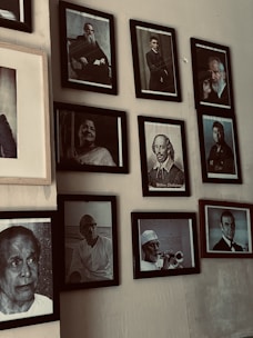 A collection of framed black and white portraits mounted on a wall. These portraits feature various individuals, some holding objects or posing formally. The frames are neatly arranged in a somewhat asymmetrical gallery layout.