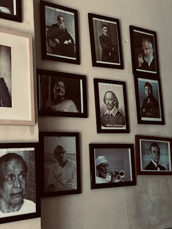 A collection of framed black and white portraits mounted on a wall. These portraits feature various individuals, some holding objects or posing formally. The frames are neatly arranged in a somewhat asymmetrical gallery layout.
