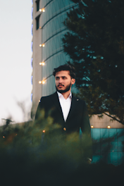 A man in a dark suit stands confidently in front of a modern glass building with illuminated edges. The background includes tall trees and the building's reflective surface, contributing to an urban atmosphere.