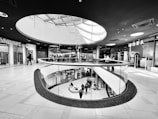 A modern shopping mall interior featuring a circular glass railing and skylight providing ample natural light. Shoppers stroll on both the upper and lower levels, surrounded by various retail stores with visible signage.