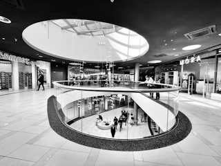A modern shopping mall interior featuring a circular glass railing and skylight providing ample natural light. Shoppers stroll on both the upper and lower levels, surrounded by various retail stores with visible signage.
