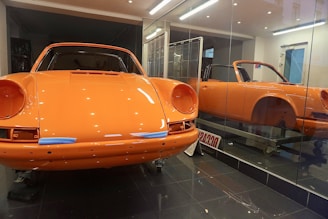 A bright orange vintage sports car is displayed inside a glass showroom. The car's body is polished and lacks some components like the headlights. There is a mirror reflection creating a duplicate image of the car. The floor is dark, and the room is well-lit, emphasizing the vibrant color of the car.