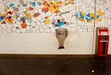 A wall with a colorful mosaic comprising various geometric shapes and vibrant colors. Below the mosaic, a white brick wall features a metallic funnel-like object with the word 'Merci' and a red fire extinguisher box labeled 'EXTINCTEUR' to its right.