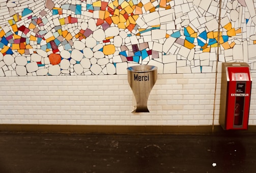A wall with a colorful mosaic comprising various geometric shapes and vibrant colors. Below the mosaic, a white brick wall features a metallic funnel-like object with the word 'Merci' and a red fire extinguisher box labeled 'EXTINCTEUR' to its right.