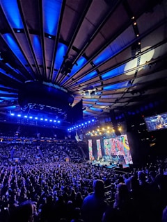 A large indoor arena filled with a crowd attending a live concert. Bright stage lights illuminate the scene, with colorful vertical screens displaying vibrant patterns. The ceiling showcases architectural beams, most prominently lit in blue hues.