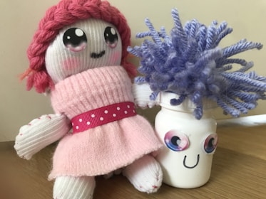 A handmade doll with pink yarn hair and a pink knitted dress sits beside a jar with a drawn smile and googly eyes, adorned with purple yarn resembling hair.