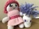 A handmade doll with pink yarn hair and a pink knitted dress sits beside a jar with a drawn smile and googly eyes, adorned with purple yarn resembling hair.