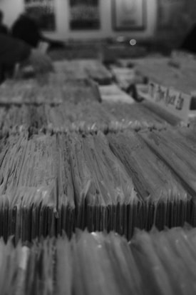 Rows of vinyl record sleeves are tightly packed in a store or collection. The focus is on the foreground with the sleeves forming neat vertical lines. In the background, though out of focus, posters or album covers are visible on the wall.