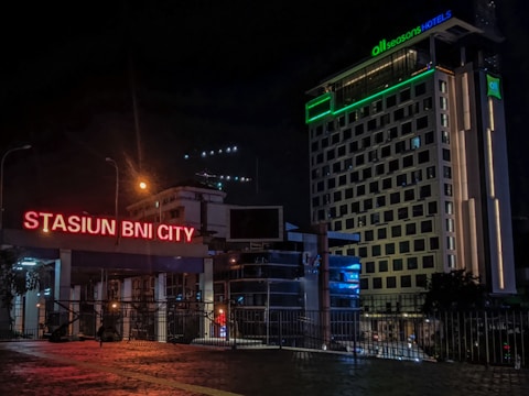 A nighttime urban scene featuring brightly lit signage for 'Stasiun BNI City' in neon red. Adjacent to the sign is a modern, multi-story hotel building with various illuminated windows and a glowing green sign for 'all seasons HOTELS'. The scene includes street lamps and the reflections of city lights on a cobblestone foreground.