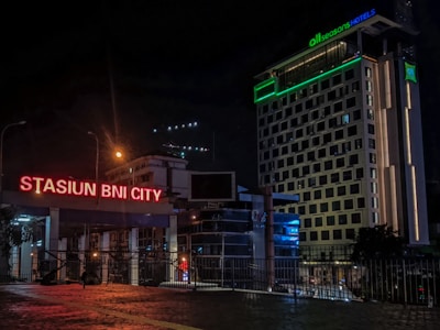 A nighttime urban scene featuring brightly lit signage for 'Stasiun BNI City' in neon red. Adjacent to the sign is a modern, multi-story hotel building with various illuminated windows and a glowing green sign for 'all seasons HOTELS'. The scene includes street lamps and the reflections of city lights on a cobblestone foreground.