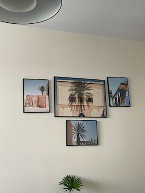 A collection of four framed photographs hangs on a light-colored wall. Each frame features different scenes, including architectural elements and palm trees. The largest photograph shows a building facade with intricate details and a palm tree in front casting shadows. The other frames include images of colorful woven textiles, additional views of palm trees, and a building corner with a blue sky backdrop. A plant with broad leaves is positioned below the frames.