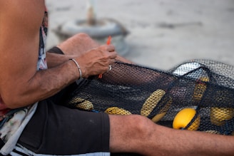 A person is sitting and working with a fishing net, threading a red tool through the netting. Yellow floats are visible within the net, indicating it is likely used for fishing. The person's arms and legs are exposed, suggesting a warm weather setting.