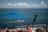 A tranquil seaside view with a large blue umbrella providing shade over a seating area. The scene includes several people relaxing, one person in orange standing by a railing watching a person on a jet ski in the water. The ocean extends far into the horizon with small boats and fluffy clouds dotting the sky.