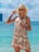 A woman stands on a beach with turquoise water in the background, wearing a light, floral summer dress and a straw hat. She has sunglasses on and is smiling, holding her hat with one hand. A small quilted bag hangs from her shoulder. The setting suggests a sunny, clear day with a rocky island visible in the distance.