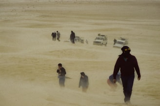 A group of people trudges through a sandstorm in a desert landscape, with four off-road vehicles partially visible in the background. The air is filled with sand, obscuring visibility and creating a harsh environment.