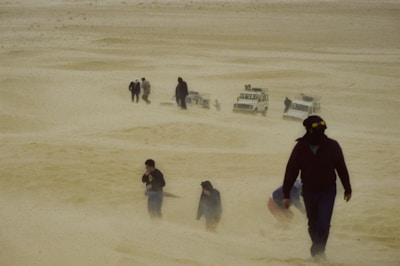 A group of people trudges through a sandstorm in a desert landscape, with four off-road vehicles partially visible in the background. The air is filled with sand, obscuring visibility and creating a harsh environment.