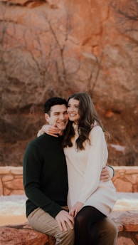A couple is sitting outdoors on a stone ledge in a natural setting with a rocky backdrop. The woman, wearing a light-colored sweater dress, has her arm gently placed around the man's neck, who is dressed in a dark green sweater. They both are smiling and appear happy.