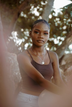 A person with tied-back hair wearing a brown tank top and a necklace with an 'M' pendant poses outdoors. The background features blurred foliage, creating a soft and natural setting.