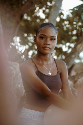 A person with tied-back hair wearing a brown tank top and a necklace with an 'M' pendant poses outdoors. The background features blurred foliage, creating a soft and natural setting.