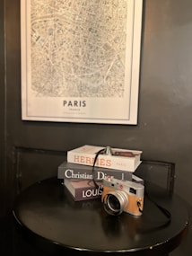 A vintage-style camera sits atop a stack of fashion brand books including 'Hermes', 'Christian Dior', and 'Louis', placed on a round black table. A detailed map of Paris is mounted on the wall behind them.
