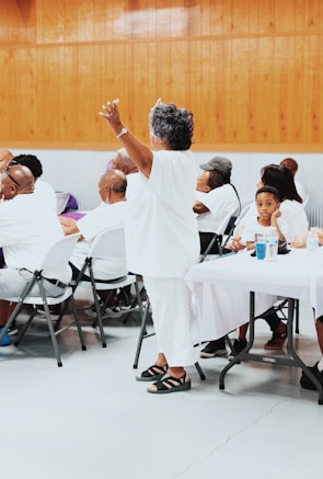 A group of people are gathered indoors, mostly seated at tables covered with white cloths. An older woman dressed in white is standing with her arms raised, surrounded by others who are listening or engaging with each other. The setting has wooden paneling on the walls and a gray floor, suggesting a community event or gathering.