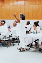 A group of people are gathered indoors, mostly seated at tables covered with white cloths. An older woman dressed in white is standing with her arms raised, surrounded by others who are listening or engaging with each other. The setting has wooden paneling on the walls and a gray floor, suggesting a community event or gathering.