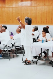 A group of people are gathered indoors, mostly seated at tables covered with white cloths. An older woman dressed in white is standing with her arms raised, surrounded by others who are listening or engaging with each other. The setting has wooden paneling on the walls and a gray floor, suggesting a community event or gathering.