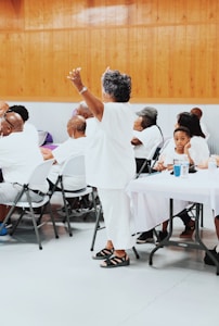 A group of people are gathered indoors, mostly seated at tables covered with white cloths. An older woman dressed in white is standing with her arms raised, surrounded by others who are listening or engaging with each other. The setting has wooden paneling on the walls and a gray floor, suggesting a community event or gathering.