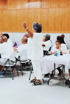 A group of people are gathered indoors, mostly seated at tables covered with white cloths. An older woman dressed in white is standing with her arms raised, surrounded by others who are listening or engaging with each other. The setting has wooden paneling on the walls and a gray floor, suggesting a community event or gathering.
