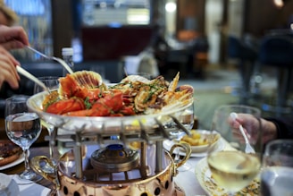 A seafood dish is served on a raised platter with various shellfish, such as lobster and clams, garnished with herbs. Surrounding the main dish are glasses of white wine and water. There are people reaching for the food with utensils, and the table setup includes plates and a decorative burner beneath the platter. The setting appears to be a restaurant with a blurred background indicating a sophisticated and cozy atmosphere.
