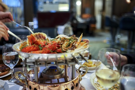 A seafood dish is served on a raised platter with various shellfish, such as lobster and clams, garnished with herbs. Surrounding the main dish are glasses of white wine and water. There are people reaching for the food with utensils, and the table setup includes plates and a decorative burner beneath the platter. The setting appears to be a restaurant with a blurred background indicating a sophisticated and cozy atmosphere.