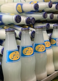 A collection of glass bottles filled with a white liquid, arranged in a staggered manner. The bottles feature a blue and yellow label with text and are capped with blue metal lids.