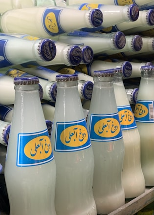 A collection of glass bottles filled with a white liquid, arranged in a staggered manner. The bottles feature a blue and yellow label with text and are capped with blue metal lids.