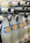 A collection of glass bottles filled with a white liquid, arranged in a staggered manner. The bottles feature a blue and yellow label with text and are capped with blue metal lids.