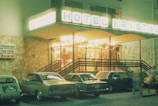 A retro-style hotel entrance with a brightly lit sign reading 'HOTEL KRISTAL'. Several vintage cars are parked in front of the stone facade. A sign on the wall advertises a restaurant, tea room, and bar. The scene is likely from the late 20th century, with the warm glow of lights creating an inviting atmosphere.