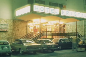 A retro-style hotel entrance with a brightly lit sign reading 'HOTEL KRISTAL'. Several vintage cars are parked in front of the stone facade. A sign on the wall advertises a restaurant, tea room, and bar. The scene is likely from the late 20th century, with the warm glow of lights creating an inviting atmosphere.