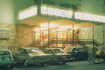A retro-style hotel entrance with a brightly lit sign reading 'HOTEL KRISTAL'. Several vintage cars are parked in front of the stone facade. A sign on the wall advertises a restaurant, tea room, and bar. The scene is likely from the late 20th century, with the warm glow of lights creating an inviting atmosphere.