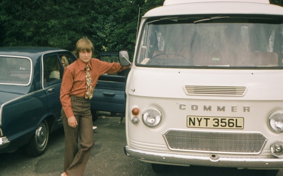 A young person leans against a vintage white Commer van with a visible license plate reading NYT 356L. Adjacent to the van is a blue car with its back door open. The person is wearing a red shirt and patterned tie, standing on an asphalt surface with greenery in the background.