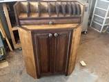A wooden cabinet with two doors and colorful knobs stands on a workshop floor. The top of the cabinet has several vertical slots for organizing or storing items. The wood has a natural finish, with darker tones on the doors and edges. Sawdust is scattered around on the concrete floor.