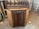 A wooden cabinet with two doors and colorful knobs stands on a workshop floor. The top of the cabinet has several vertical slots for organizing or storing items. The wood has a natural finish, with darker tones on the doors and edges. Sawdust is scattered around on the concrete floor.