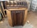 A wooden cabinet with two doors and colorful knobs stands on a workshop floor. The top of the cabinet has several vertical slots for organizing or storing items. The wood has a natural finish, with darker tones on the doors and edges. Sawdust is scattered around on the concrete floor.