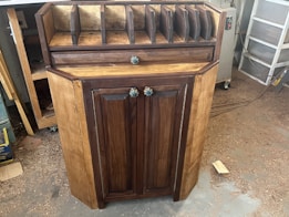 A wooden cabinet with two doors and colorful knobs stands on a workshop floor. The top of the cabinet has several vertical slots for organizing or storing items. The wood has a natural finish, with darker tones on the doors and edges. Sawdust is scattered around on the concrete floor.