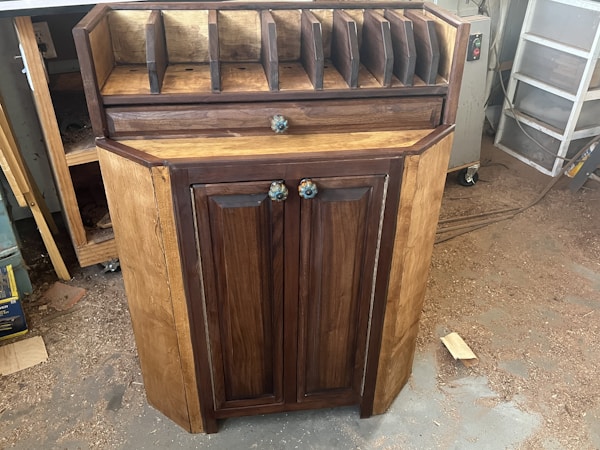 A wooden cabinet with two doors and colorful knobs stands on a workshop floor. The top of the cabinet has several vertical slots for organizing or storing items. The wood has a natural finish, with darker tones on the doors and edges. Sawdust is scattered around on the concrete floor.