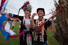 Two people are dressed in vibrant traditional outfits with intricate patterns and colorful beadwork. One person wears a headpiece adorned with multicolored pom-poms, while the other holds a traditional musical instrument. They appear to be in a festive outdoor setting, with grass and trees in the background.