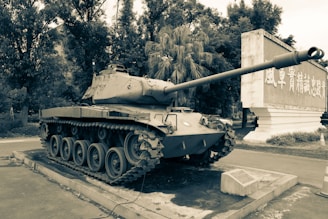 A vintage military tank is displayed on a concrete pedestal, surrounded by trees and accompanied by a stone plaque. The scene is outdoors, with a large sign featuring Chinese characters in the background.