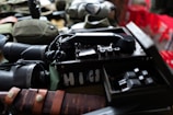 A collection of vintage military items including a field radio with a rotary dial, a pair of binoculars, and a knife with a leather sheath. The objects are arranged on a surface, with some additional gear partially visible in the background.